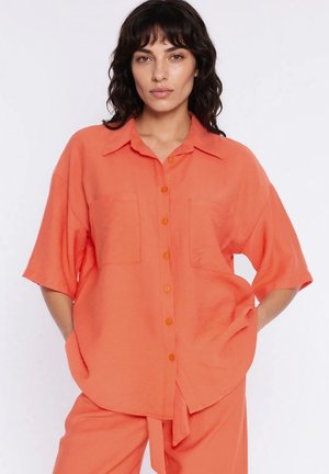 Kobieta o ciemnych, kręconych włosach, ubrana w luźną pomarańczową koszulę z krótkim rękawem i pasujące spodnie, stojąca z rękami w kieszeniach.