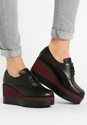 Czarne skórzane buty na platformie z burgundowymi akcentami na podeszwach, noszone z podwiniętymi jasnoszarymi dżinsami na białym tle.