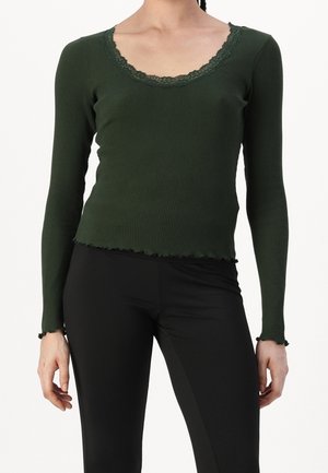 Frau trägt ein dunkelgrünes, geripptes Langarmshirt mit Spitzenbesatz, kombiniert mit schwarzer, enger Hose vor einfachem Hintergrund.