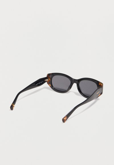 Γυαλιά ηλίου σε σχήμα ματιών γάτας με μαύρο και χελώνα πλαίσιο και σκούρους φακούς, τοποθετημένα πάνω σε λευκή επιφάνεια, με γωνία που δείχνει το μπροστινό μέρος και τα μπράτσα.