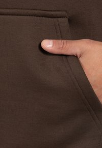 Brązowy materiał swetra z prążkowanym teksturą. Dłoń jest umiejscowiona w pobliżu kieszeni, podkreślając detale szwów i szwu kieszeni.