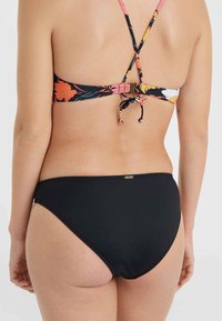 Bikini komplekts ar ziedu rakstu augšdaļu, kas aprīkota ar regulējamiem melniem siksniņām, un vienkrāsainām melnām apakšdaļām ar zelta logo detaļu.