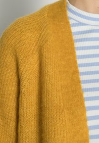 Hořčičně žlutý pletený cardigan přes bílou a světle modrou pruhovanou ribbed košili, odhalující ramena a oblast výstřihu.