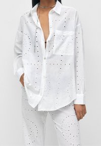 Chemise blanche en coton à boutons avec des motifs découpés et une poche poitrine, associée à un pantalon assorti présentant des détails découpés similaires.