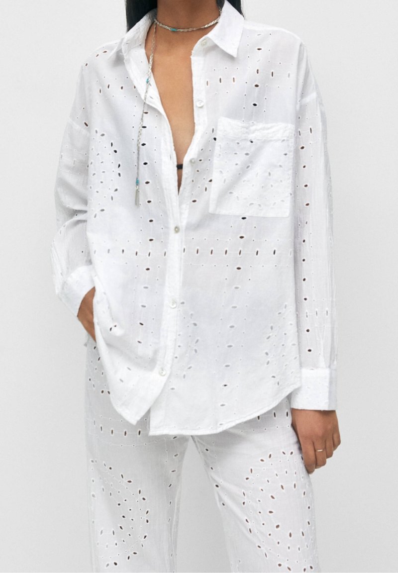 Chemise blanche en coton à boutons avec des motifs découpés et une poche poitrine, associée à un pantalon assorti présentant des détails découpés similaires.