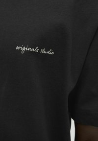 Sort t-shirt lavet af bomuld, med en lille hvid broderet tekst "originals studio" på venstre forsideside.