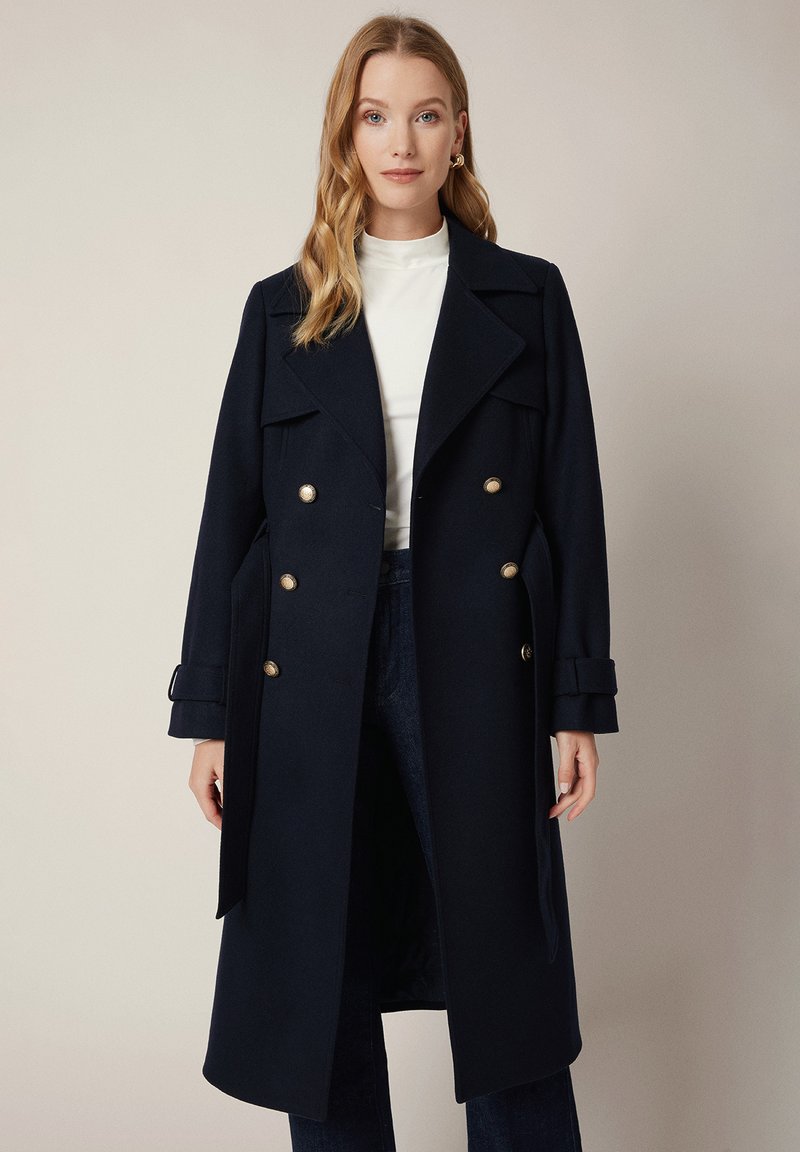 Granatowy płaszcz dwurzędowy wykonany z wełny, z ozdobnymi złotymi guzikami, szerokimi klapami i paskami u mankietów. Gładka tekstura i dopasowany krój.