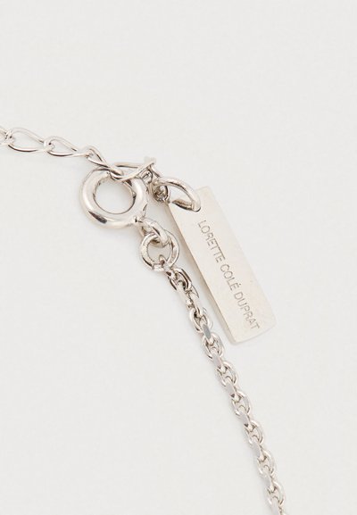 Sidabrinės grandinėlės užsegimas su apvaliu spyruokliniu žiedu ir stačiakampiu žymekliu, kuriame įrėžtas užrašas „LORETTE COLE DUPRAT“ ant balto fono.