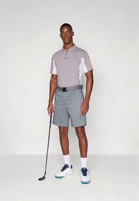 Γκρι πόλο πουκάμισο με λευκές λεπτομέρειες, γκρι σορτς με καρό σχέδιο και μαύρη ζώνη. Στενή εφαρμογή· το ύφασμα φαίνεται να απορροφά την υγρασία. Μαύρο μπαστούνι γκολφ κρατημένο στο χέρι.