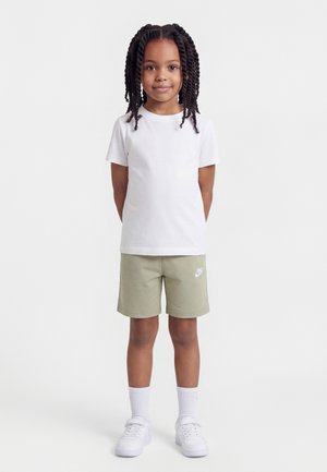 Niña joven con trenzas, vestida con camiseta blanca, pantalones cortos verdes claros de Nike, calcetines blancos y zapatillas blancas, de pie con las manos detrás de la espalda.