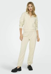 Felpa zip-up color crema con polsini a coste, abbinata a pantaloni jogger coordinati. Il modello indossa mocassini neri. Tessuto morbido, design casual.