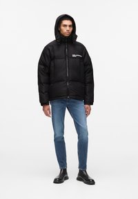 Sort pufferjakke med lynlås og hætte, med et logo på brystet; bæres med blå jeans og sorte støvler.