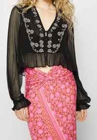 Blusa nera trasparente con motivi floreali ricamati, orlo arricciato e chiusura con bottoni. Abbinata a una gonna avvolgente rosa con fiori e dettagli.