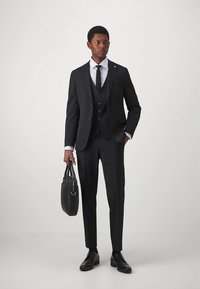 Zwart driedelig pak met een getailleerd colbert, gilet en broek. Gecombineerd met een wit overhemd, zwarte stropdas en leren tas. Klassiek ontwerp en pasvorm.