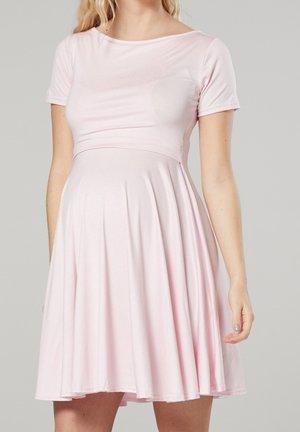 Femme portant une robe rose pâle à manches courtes, longueur genou, avec un corsage ajusté et une jupe évasée, sur un fond uni.