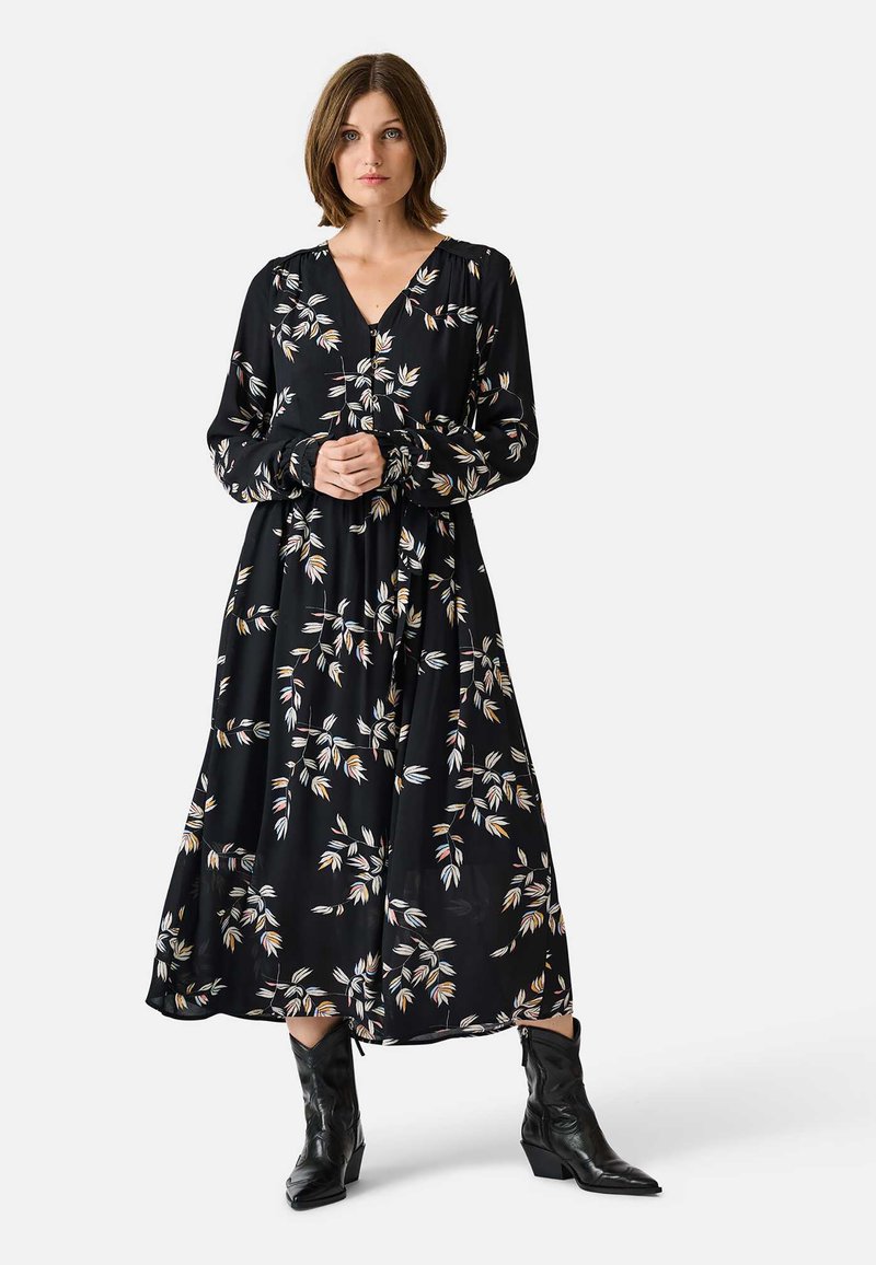 Black floral dress with long sleeves, V-neck, and a flowing silhouette. Features small leaf patterns in white, yellow, and green. Worn with black boots.