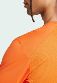 Camiseta deportiva naranja con tela suave y paneles de malla texturizados en los hombros. Vista en primer plano de la espalda y el escote.