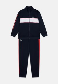 Survêtement marine avec bande blanche sur la poitrine et accents rouges. Veste zippée avec col montant ; le pantalon présente des bandes sur les côtés. Logo Lacoste affiché.