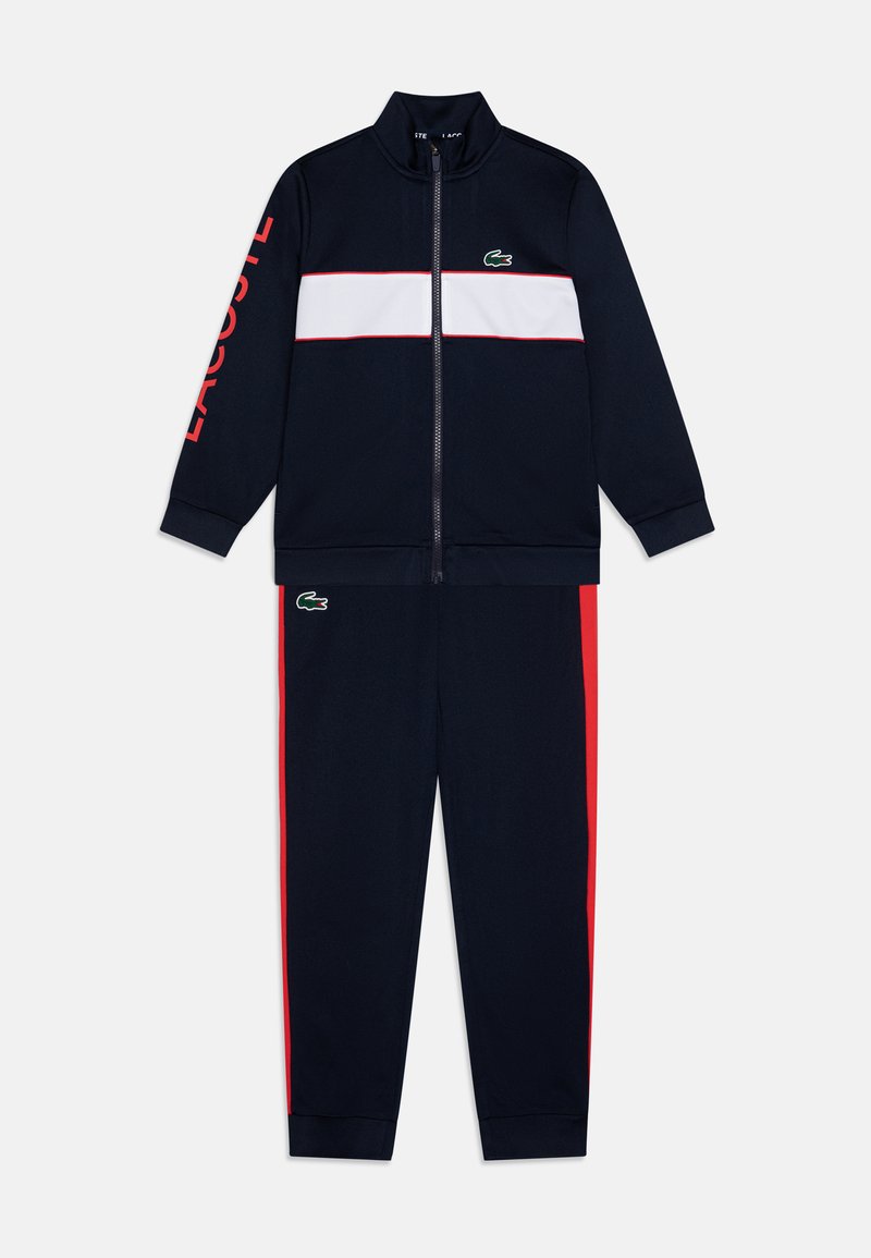 Fato de treino azul-marinho com faixa branca no peito e detalhes em vermelho. Casaco com fecho éclair e gola alta; as calças têm faixas laterais. Logotipo da Lacoste exibido.