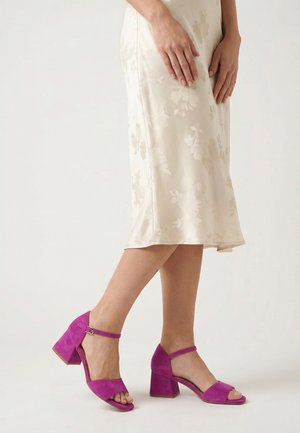 Kvinde iført en cremefarvet blomsterprint midi-nederdel og lyserøde ruskind block-hælede sandaler med ankelremme, stående på en lys baggrund.
