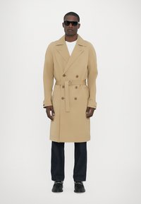 Trench coat dublu cu bucăți, de culoare bej, cu talie reglabilă, rever în formă de notch și buzunare laterale. Asortat cu blugi închiși la culoare și pantofi negri.