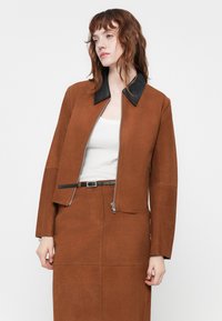 Brązowa zamszowa kurtka typu cropped z czarnym skórzanym kołnierzem, zamek z przodu i długimi rękawami; noszona na białej bluzce i pasującej brązowej spódnicy.