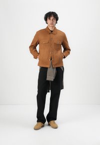 Bruine suède jas met twee borstzakken, gecombineerd met een grijs geruit overhemd en zwarte broek. Model draagt beige sneakers. Eenvoudig, casual ontwerp.