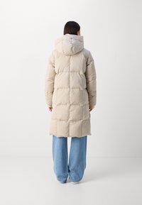 Beige pufferjas met een capuchon, voorzien van een gewatteerd ontwerp en een middellange snit, gecombineerd met lichtblauwe wijde jeans.
