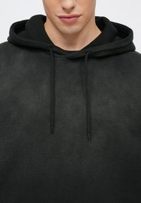 Μαύρο φούτερ με κουκούλα και απαλή υφή, με κορδόνια που έχουν πλαστικές άκρες και χαλαρή εφαρμογή. Χωρίς σχέδια ή λογότυπα ορατά.