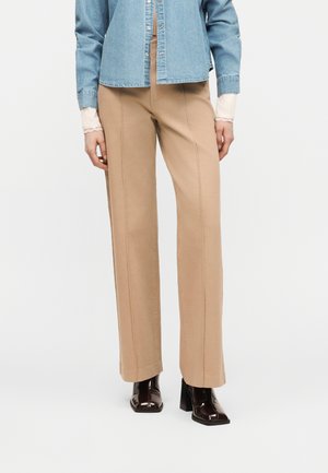 Kvinde iført beige bukser med lige ben, blå denimjakke, hvid top med blonder ved manchetterne og mørkebrune støvler med firkantet tå og hæl.