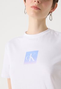 Tricou scurt cu mânecă albă, prezentând un pătrat degradat în nuanțe de albastru și violet, cu logo-ul "cK" imprimat pe piept. Textile din bumbac neted.