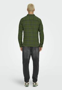 Zielona koszula w kratę, wykonana z materiału, z czarnymi i białymi liniami tworzącymi siatkę, z guzikami przy mankietach oraz prostym dołem. Noszona z ciemnymi dżinsami i trampkami.