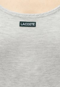 Gros plan d’un tissu gris clair avec une étiquette rectangulaire vert foncé portant l’inscription « LACOSTE », cousue près de l’encolure.