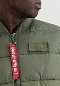 Olivgrüne Bomberjacke mit gesteppter Textur, geripptem Kragen und einem Patch-Logo. Enthält ein rotes "Remove Before Flight"-Tag.