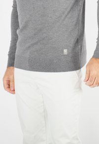 Pull gris en laine douce, avec un ourlet et des poignets côtelés, orné d'un petit badge logo sur le devant en bas. Porté avec un pantalon blanc.