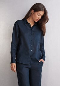 Camicia di lino blu navy con maniche lunghe, colletto, tasca frontale e patta di bottoni. Tessuto morbido e vestibilità comoda. Abbinata a pantaloni coordinati.