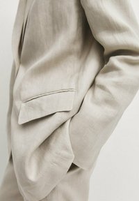 Gros plan sur la poche et la manche d'une veste de costume en lin beige, avec une main glissée négligemment dans la poche.