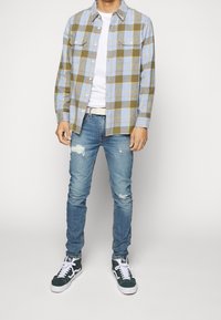Camicia di flanella a quadri in azzurro chiaro e oliva con due tasche sul petto, indossata sopra una maglietta bianca. Jeans blu strappati e sneakers verdi.