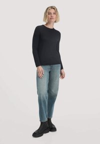 Frau mit kurzen blonden Haaren, die ein schwarzes Langarmoberteil, hellblaue Jeans und schwarze Stiefeletten trägt, steht vor einem einfarbigen Hintergrund.