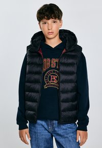 Schwarze, gepolsterte Weste mit Kapuze, Reißverschluss und gesteppter Textur, getragen über einem marineblauen Hoodie mit grafischem Druck. Legere blaue Jeans.
