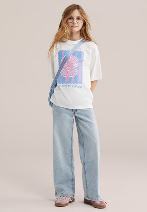 Jong persoon met lang haar, een ronde bril, een wit T-shirt met een schelpafbeelding en tekst, lichtblauwe spijkerbroek, roze schoenen en een schoudertas.