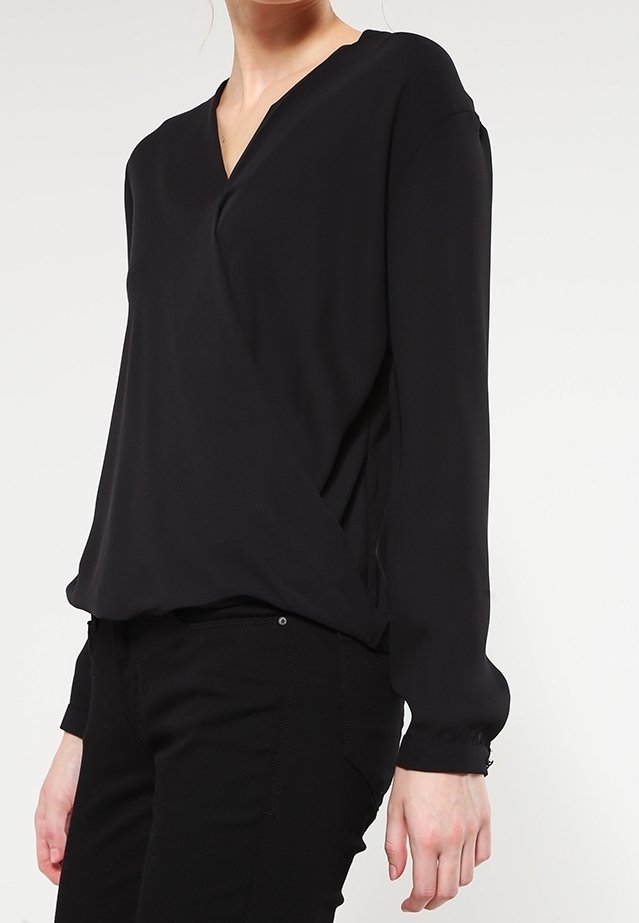 Femme portant un chemisier noir à manches longues avec un col en V, assorti à un pantalon noir, sur un fond blanc uni.