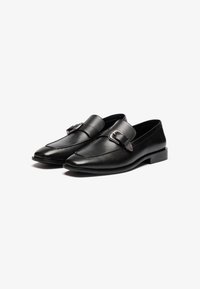 Mocasines de cuero negro con punta afilada, que presentan una hebilla decorativa y una textura suave. Tacón bajo y diseño elegante.