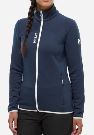 Femme aux cheveux tressés portant une veste zippée bleu marine avec bordure blanche, poches zippées, et logos "MILLET" sur la poitrine et la manche.