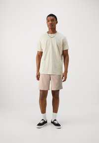 Camiseta de algodón beige, de manga corta, combinada con pantalones cortos a cuadros en colores crema y óxido. El modelo lleva zapatillas negras y calcetines blancos.