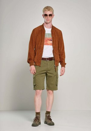 Braune Wildleder-Bomberjacke, weißes T-Shirt mit Grafik, olivgrüne Cargo-Shorts, brauer Gürtel, braune Stiefel, Sonnenbrille, heller Hintergrund.