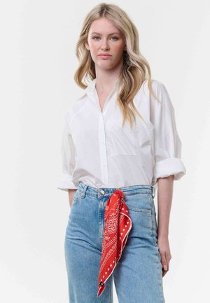 Junge Frau mit langen blonden Haaren trägt ein weißes Hemd mit Knöpfen, hellblaue Jeans und ein rotes, gemustertes Bandana, das an den Jeans befestigt ist.