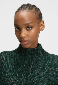 Mörkgrön stickad tröja med hög krage. Har ett texturerat ribbmönster och subtila färgvariationer för djup.