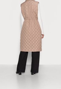 Gilet matelassé long sans manches en beige clair, avec une taille cintrée et un motif en diamant, associé à un pantalon large noir et des talons épais.