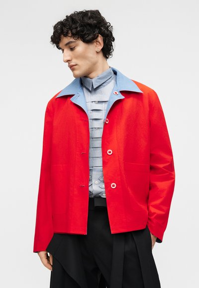 Νέος άνδρας με σγουρά μαλλιά φοράει ένα φωτεινό κόκκινο μπουφάν με γαλάζιο γιακά πάνω από μια πλισέ γκρι πουκαμίσα και άνετο μαύρο παντελόνι.